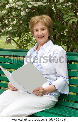 Elderly woman with a illustrated magazine in hands. Sits on a bench in a garden Smiles and looks in a photographic camera