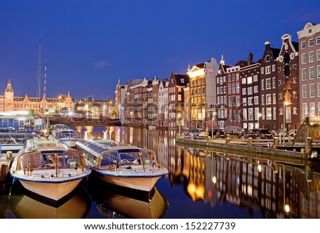 City of Amsterdam in Netherlands at night, historic apartment houses with reflections on water and boats ready for canal tours and cruises.