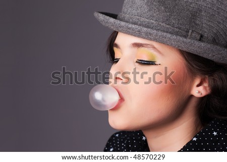 Closeup of a pretty young girl with professional makeup blowing bubble gum against gray background.