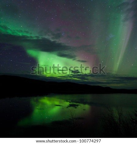 Night Sky Stars, clouds and Northern Lights mirrored on calm lake in Yukon, Territory, Canada.