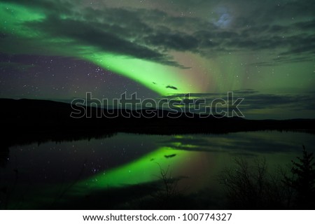 Night Sky Stars, clouds and Northern Lights mirrored on calm lake in Yukon, Territory, Canada.