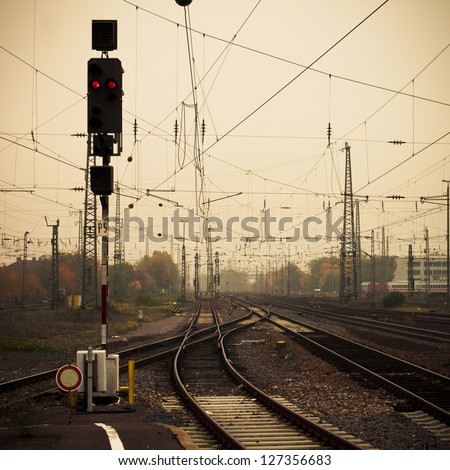 Moble photography lo-fi styled image of confusing urban railway tracks with ines and overhead cables and a red signal light at foggy dusk