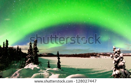 Spectacular display of intense Northern Lights or Aurora borealis or polar lights forming green arc over snowy winter landscape