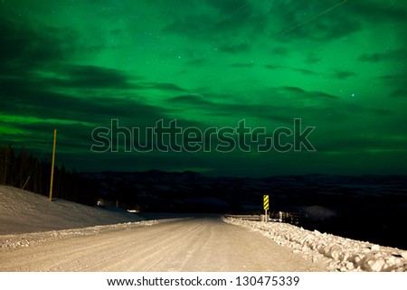 Northern Lights or Aurora borealis or polar lights illuminating overcast sky over snowy winter road landscape