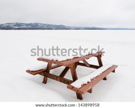 Closed for the season: beautiful lakeside campsite with wooden table and benches burried in snow and wide open flat of snowy frozen lake landscape with pristine white snow on a cold winters day