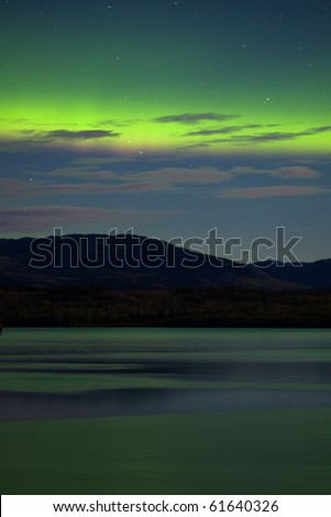 Intense Aurora borealis (northern lights) showing between clouds during moon lit night being mirrored on Lake Laberge, Yukon Territory, Canada.
