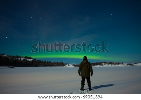 Man watching Northern Lights (Aurora borealis) over moon lit snowscape of frozen lake and forested hills.