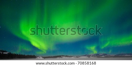 Northern Lights (Aurora borealis) over moon lit snowscape of frozen lake and forested hills.