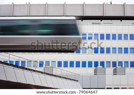 Modern public transportation system sky train hanging from elevated guide way  passes by in front of tall office building facade (with motion blur).