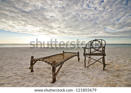 Old Bamboo Chair and Camp Bed Sitting on Sandy Beach with Sea and Setting Sun Over Dramatic Sky.
