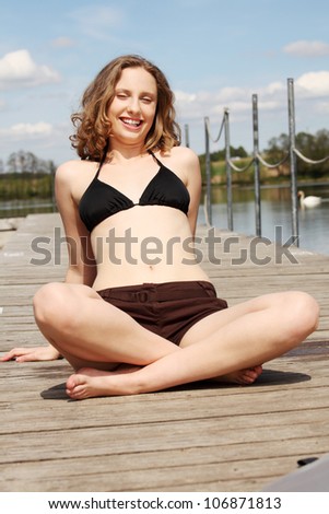 Young woman is sitting with crossed legs on the bridge in sunny day. Pretty girl in bikini is laughing.