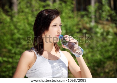 Young woman during warm, sunny day, drinking water to cool down. Pretty girl holding bottle of cold water next to her lips and pouring drink to her mouth.