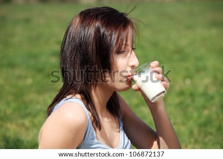 Young woman during warm, sunny day, wearing summer dress, drinking milk. Pretty girl on meadow is holding cup (glass).