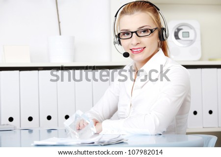 Young woman sitting at desk in office with headphones set working in call center. Blond secretary/receptionist in glasses and white shirt talking through microphone as help desk consultant.