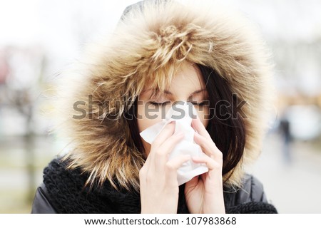 Young woman wearing furry hood, sneezes during cold day. Woman is holding tissue next to her nose.