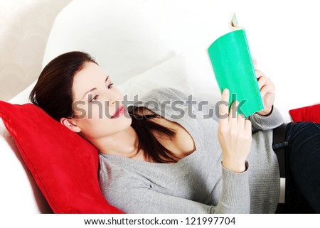 Portrait of a beautiful young woman resting her head on a red pillow, lying on a sofa and reading a book.