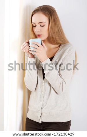 Beautiful casual caucasian woman standing by the window with hot drink coffee or tea. Indoor background.