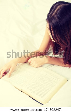 Beautiful young brunette woman reading a book on the bed, at home