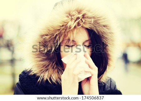 Young woman wearing furry hood, sneezes during cold day. Woman is holding tissue next to her nose.