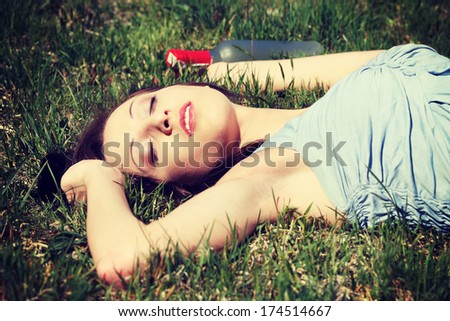 Drunk young woman is sleeping on the grass next to bottle of alcohol in sunny day.