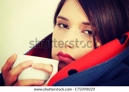 Young woman caught cold, wrapped up in blanket, drinking something hot from cup.