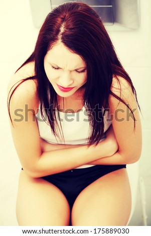 Front view of a suffering woman because of the belly ache, sitting in bathroom embracing her stomach.