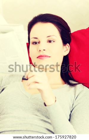 Front view portrait of a young beautiful smiling woman lying on a sofa, resting her head on a red pillow and reading a neswpaper.