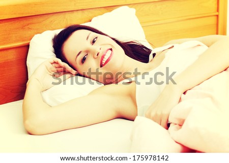 Closeup of a young beautiful woman resting in the bed, holding her right hand next to the face and smiling to the camera.