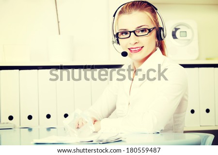Young woman sitting at desk in office with headphones set working in call center. Blond secretary/receptionist in glasses and white shirt talking through microphone as help desk consultant.