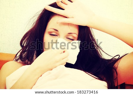 Front view face closeup of a beautiful young woman lying in bed, having a cold, sneezing in a tissue and resting her hand on the forhead.
