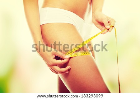 Closeup photo of a Caucasian woman\'s leg. She is measuring her thigh with a yellow metric tape measure after a diet
