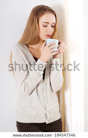 Beautiful casual caucasian woman standing by the window with hot drink coffee or tea. Indoor background.