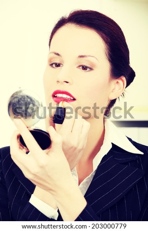 Beautiful business woman is putting lipstick on her lips and holding mirror in office.