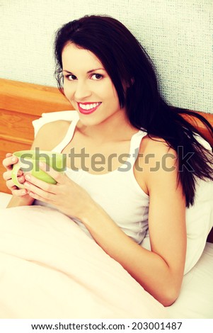 Smiling woman is holding cup of coffee or tea and sitting in bed, in home.