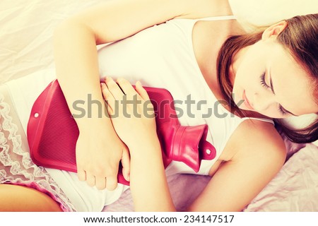 Beautiful young woman is lying on the bed with hot water bottle, have stomach ache.