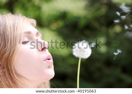 The blonde sits in park on a grass with a dandelion in hands and blowing