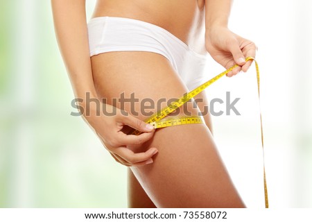 Closeup photo of a Caucasian woman\'s leg. She is measuring her thigh with a yellow metric tape measure after a diet