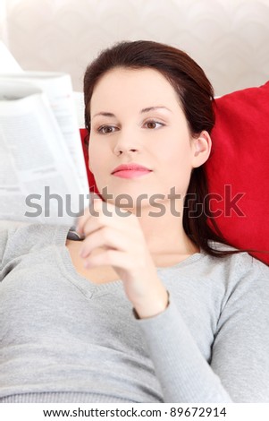 Front view portrait of a young beautiful smiling woman lying on a sofa, resting her head on a red pillow and reading a neswspaper.