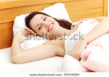 Closeup of a young beautiful woman resting in the bed, holding her right hand next to the face and smiling to the camera.