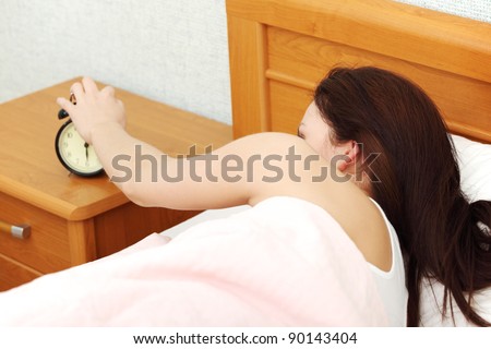Back view closeup of a young beautiful woman reaching out her hand to turn off a black alarm clock.