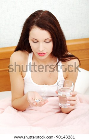 Front view portrait of a beautiful young woman in bed, holding pills and a glass of water.