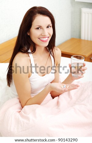 Portrait of a beautiful young woman in bed, holding pills and a glass of water, smiling to the camera.