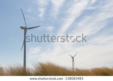 Two wind turbines against a blue sky towering over the grass covered dunes.