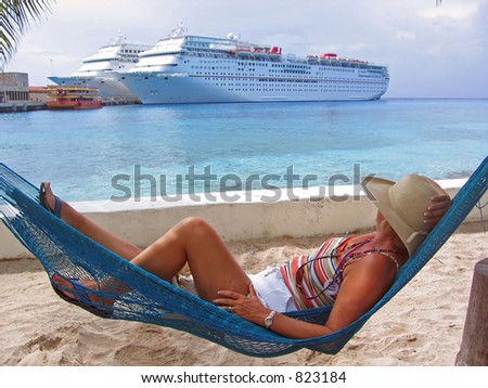 A woman lies in a hammock on a sandy beach and looks over at cruise ships in the port
