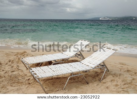 Two empty lounge chairs on an island beach with a cruise ship in the background