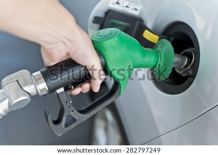 Close up of filling car with petrol. Image shows male hand holding fuel pump having a firm grip and filling a silver coloured car. Shallow focus on fuel pump. Pump is black and green.