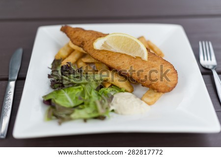 Piece of crumbed fish placed on top of chips with a slice of lemon on top. Garden salad and tartar sauce on side. Square plate on outdoor wooden table.