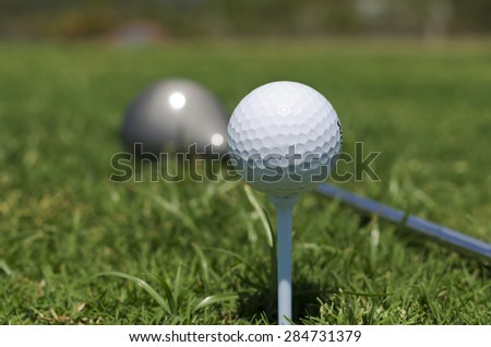 Close up shot of a white golf ball sitting on a tee. Long green grass around and out of focus golf club in background.