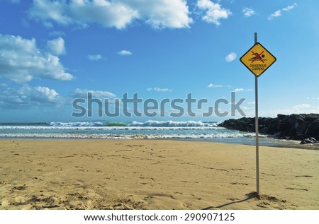 Dangerous current sign on beach. Diamond shaped orange or yellow sign with figure of swimmer crossed out with red. Sign attached to a pole and stuck in the sand on a beach.