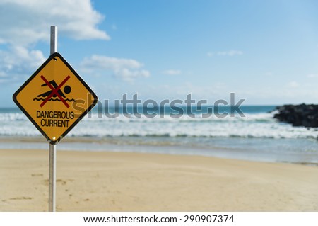 Dangerous current sign on beach. Diamond shaped orange or yellow sign with figure of swimmer crossed out with red. Sign attached to a pole and stuck in the sand on a beach. Selected focus on sign.